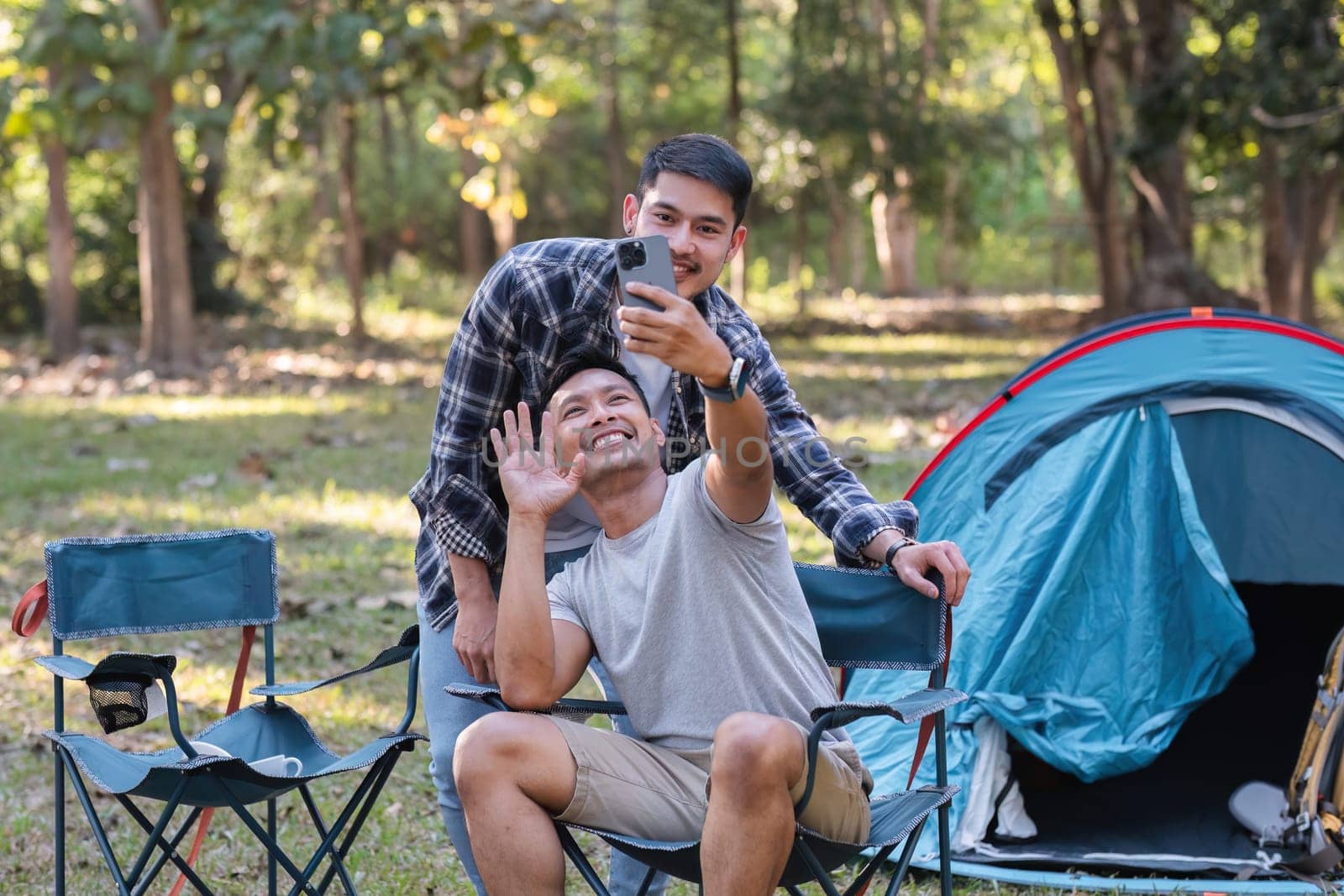 Asian LGBTQ couple tent in front of camp talking on smartphone in video chat or take a selfie by wichayada