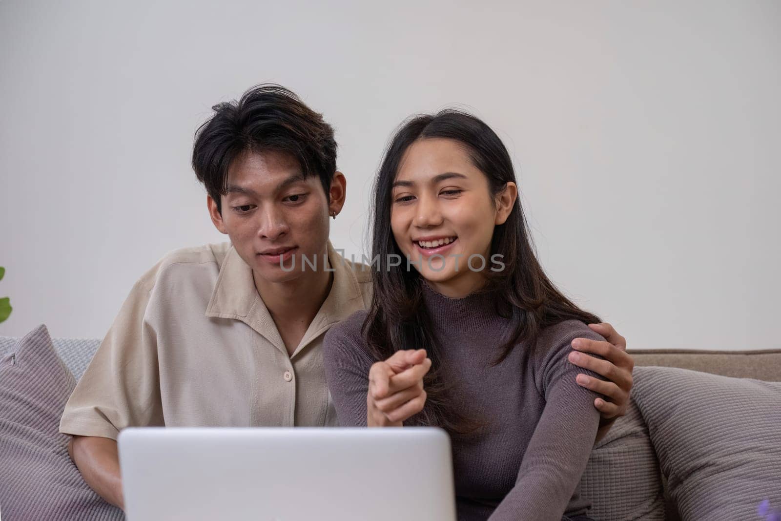 Asian couple watching movies on the internet using laptop at home Smiling Thai man and woman sitting on sofa, hugging each other, looking at computer screen. Surf the web together copy space.