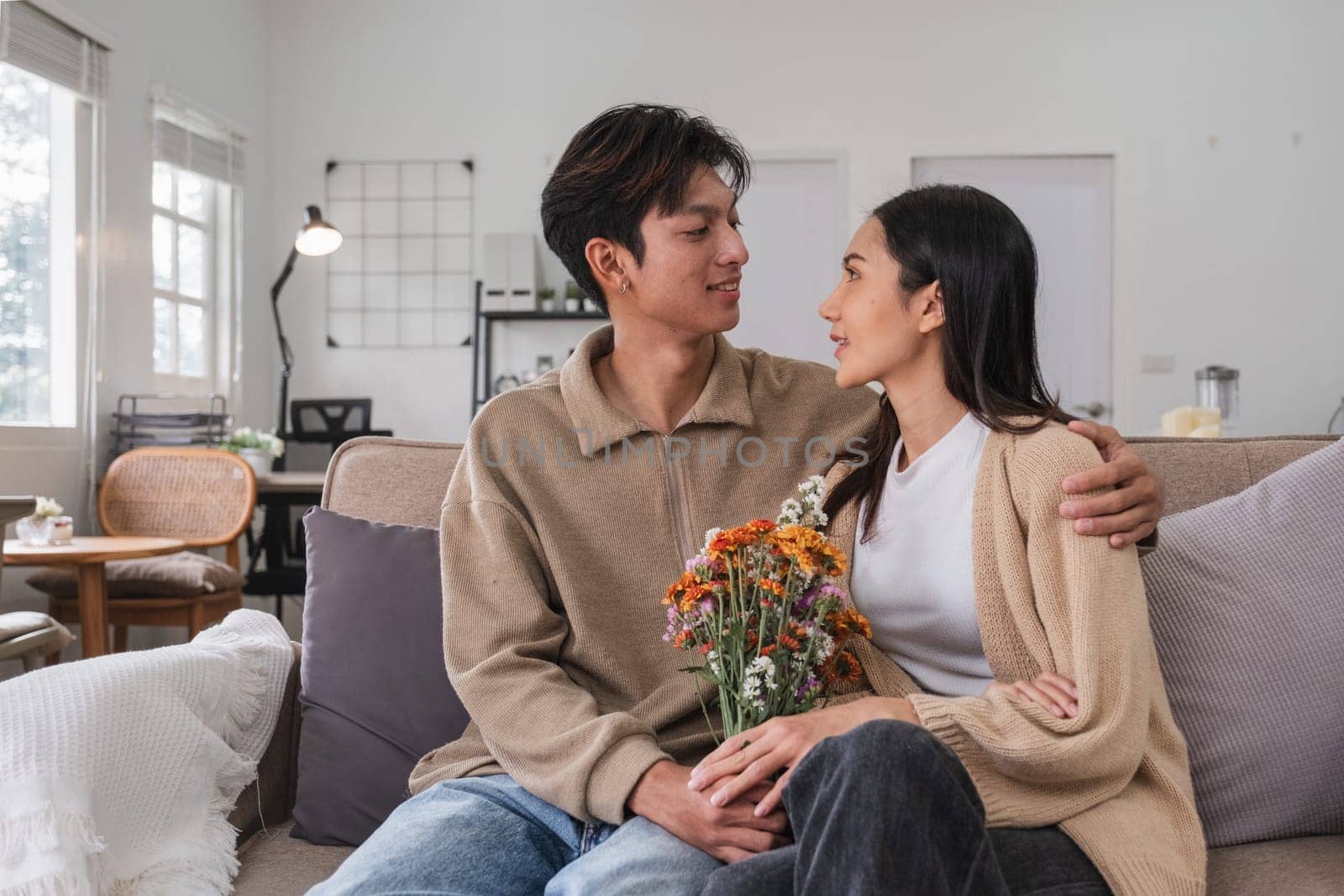 Romantic young asian couple embracing with holding flowers and smiling in living room at home. fall in love. Valentine concept by wichayada