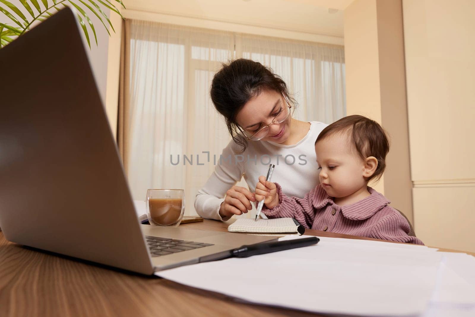 Cheerful pretty businesswoman working on laptop at home with her little child girl. mom spending time with her cute baby