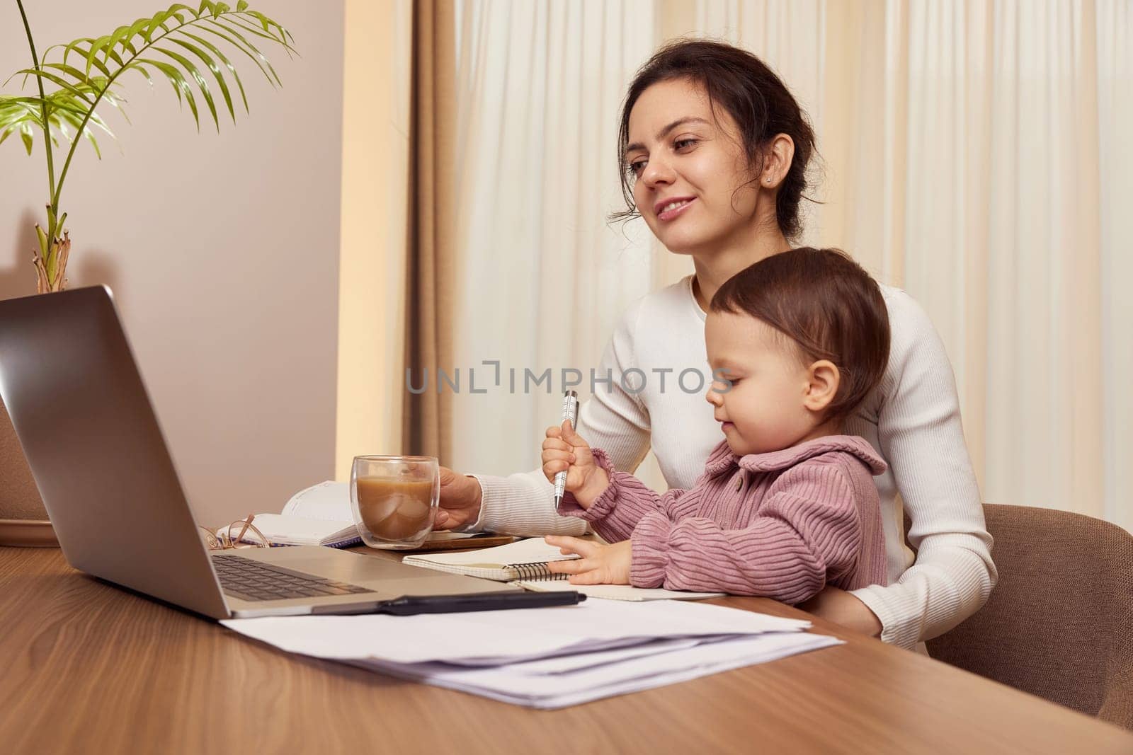 Cheerful pretty businesswoman working on laptop at home with her little child girl. mom spending time with her cute baby