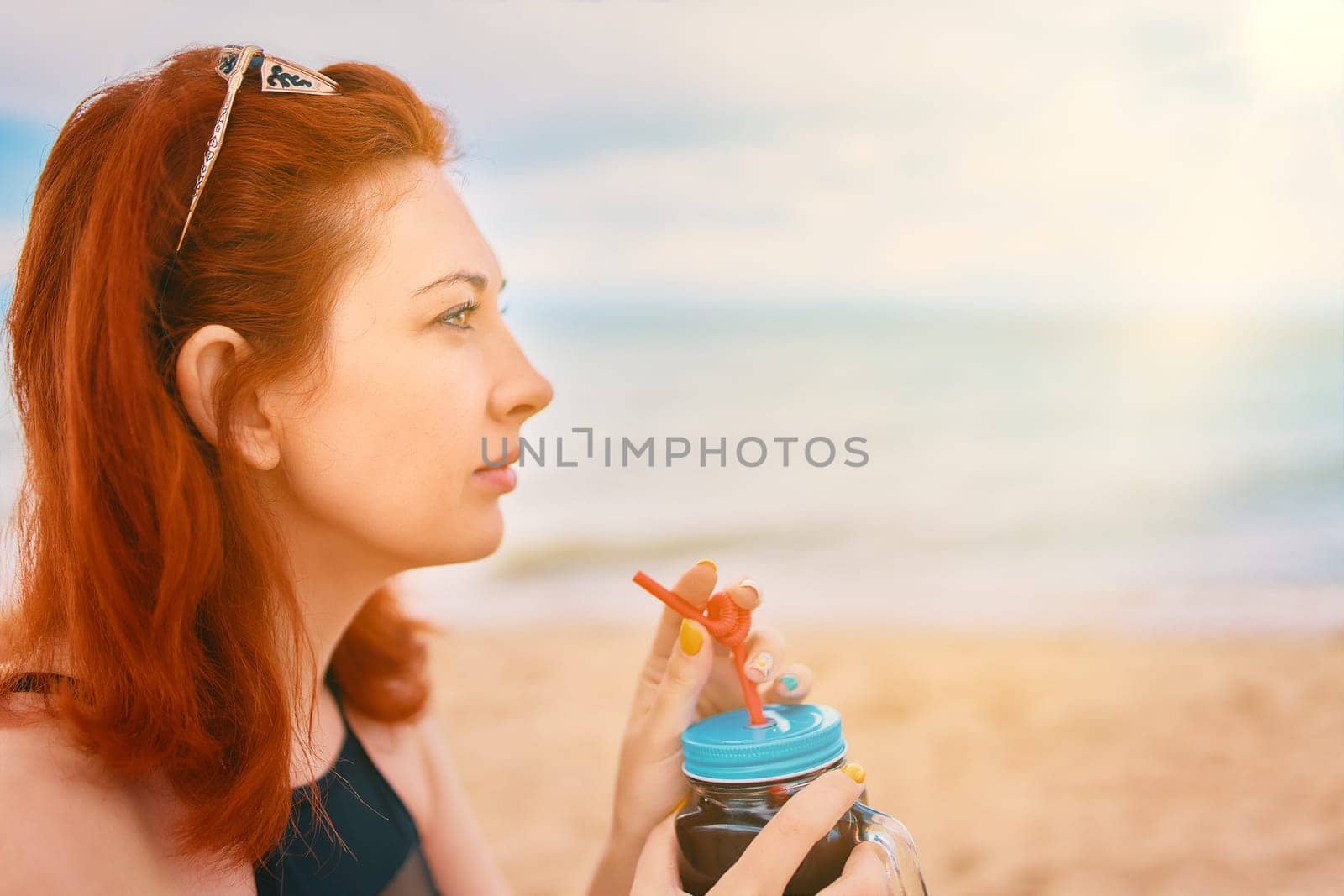 Red-haired girl in a swimsuit is sitting on the beach with a cocktail and look away. Calm woman on vacation. Holidays at the sea. Vintage sunglasses on the head of a having a rest lady.