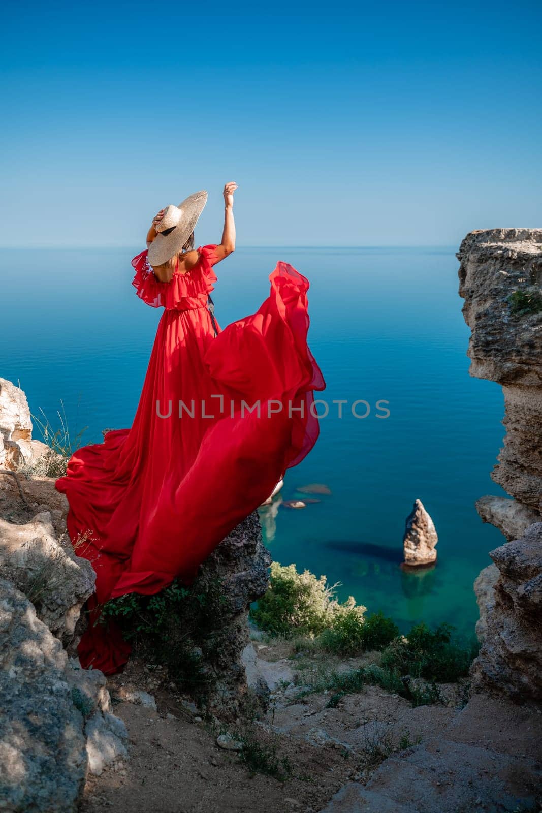 Red dress sea woman. Happy woman with flowing hair in a long flowing red dress stands on a rock near the sea. Travel concept, photo session at sea by Matiunina