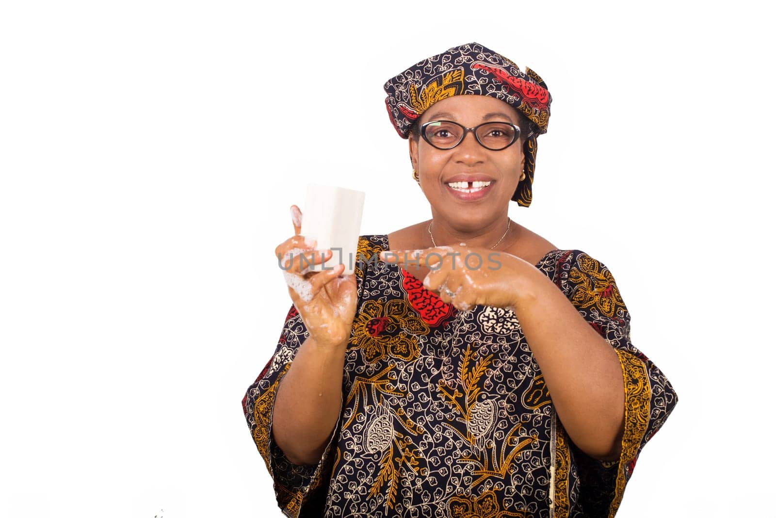 Happy woman dressed in african outfit with scarf on head holds and presents white soap to camera with hand gesture isolated on white background.