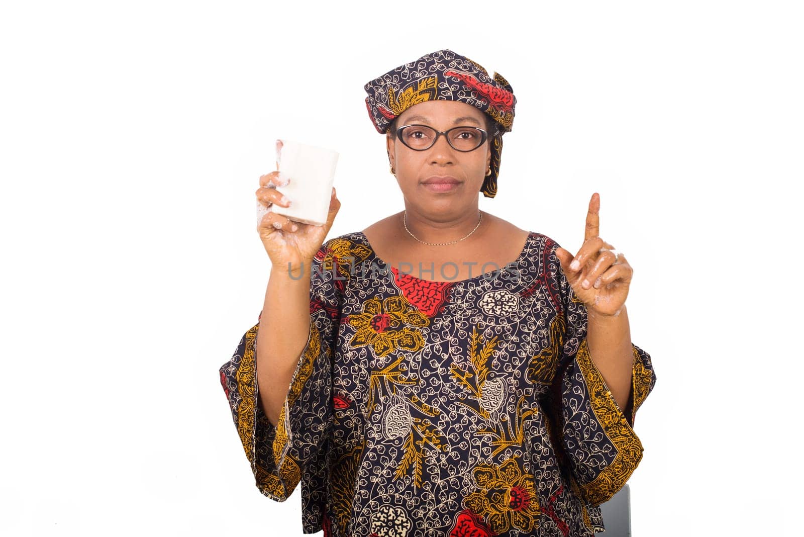 Happy woman dressed in african outfit with scarf on head holds and presents white soap to camera with hand gesture isolated on white background.