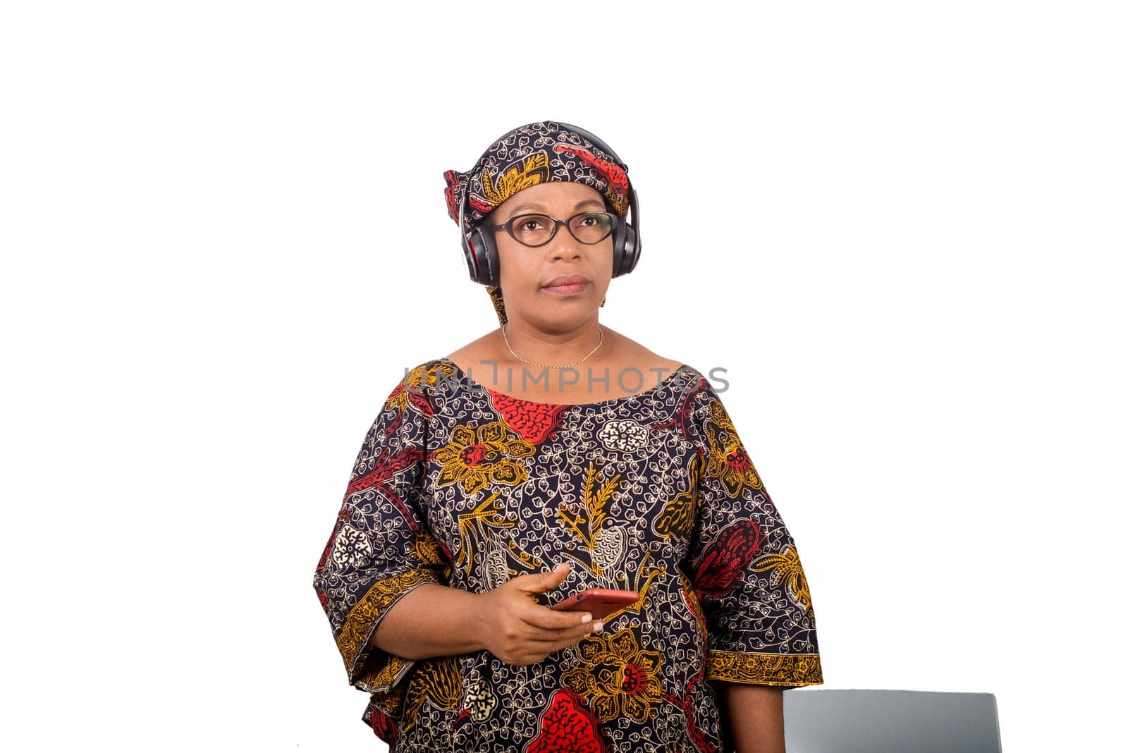 smiling mature african woman in african loincloth and scarf listening music with headphones on a cellphone over white background. she looks up and thinks of something