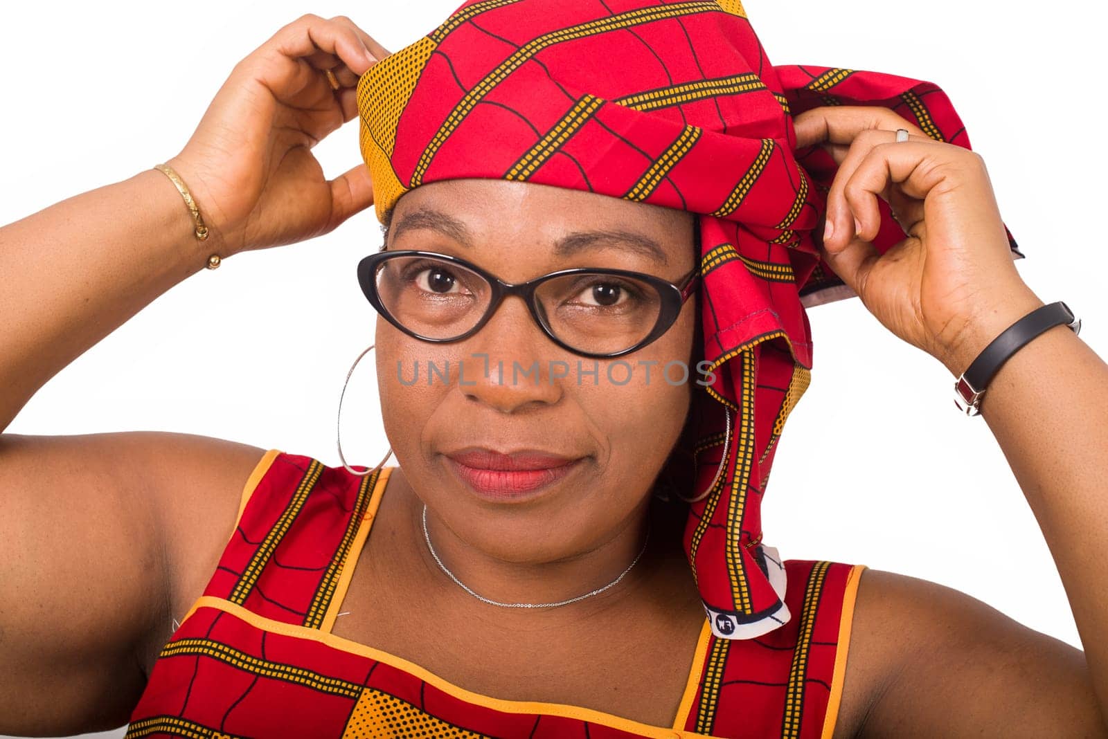 Close-up stylish smiling african mature woman in red african loincloth and glasses standing in studio and arranging her traditional headscarf on her head