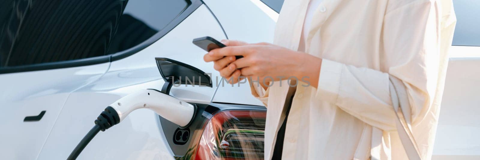 Young woman use smartphone to pay for electricity at public EV car charging station green city park. Modern environmental and sustainable urban lifestyle with EV vehicle. Panorama Expedient