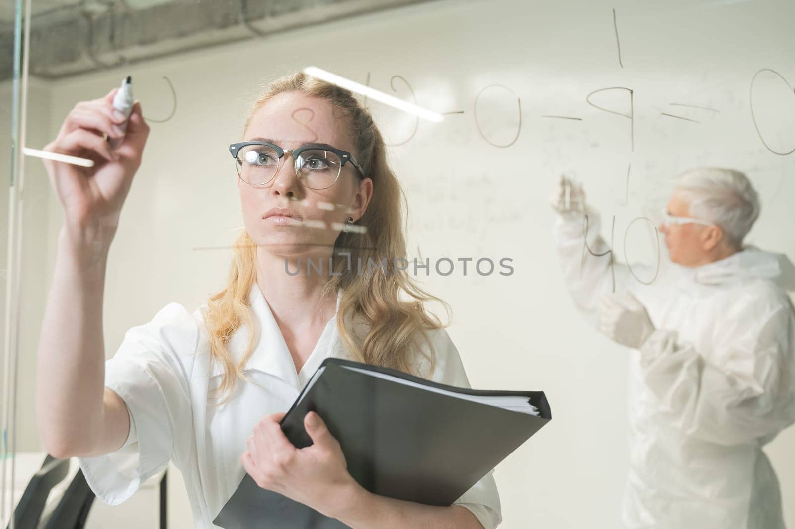 A woman chemist writes a formula on glass. An elderly Caucasian man in a protective suit is doing tests