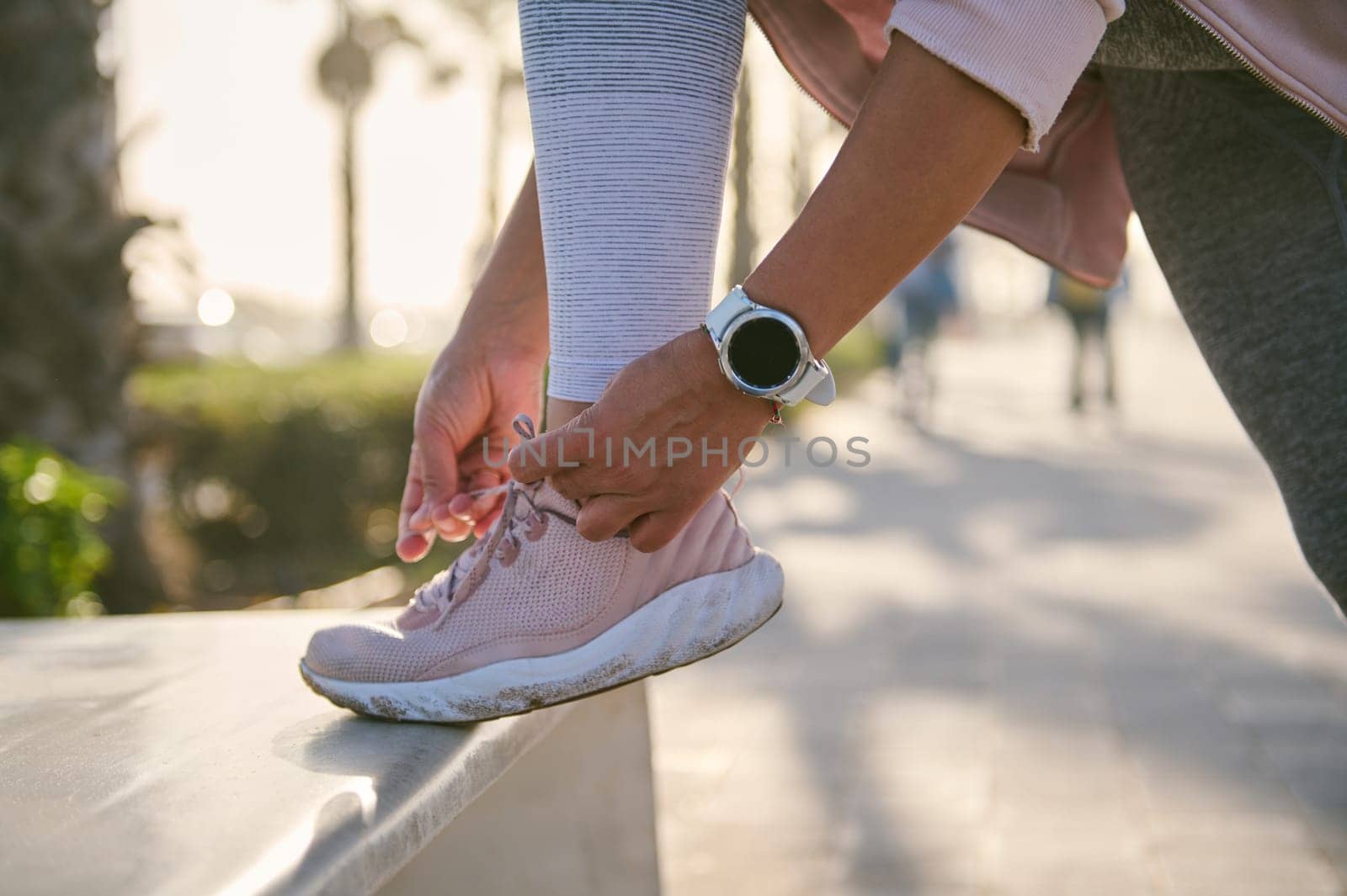 Cropped view of a female athlete, sportswoman tying laces on pink sports shoes, wearing smart wristwatch on hand for checking and pulse control, heart rate and loosed calories, ready for morning jog