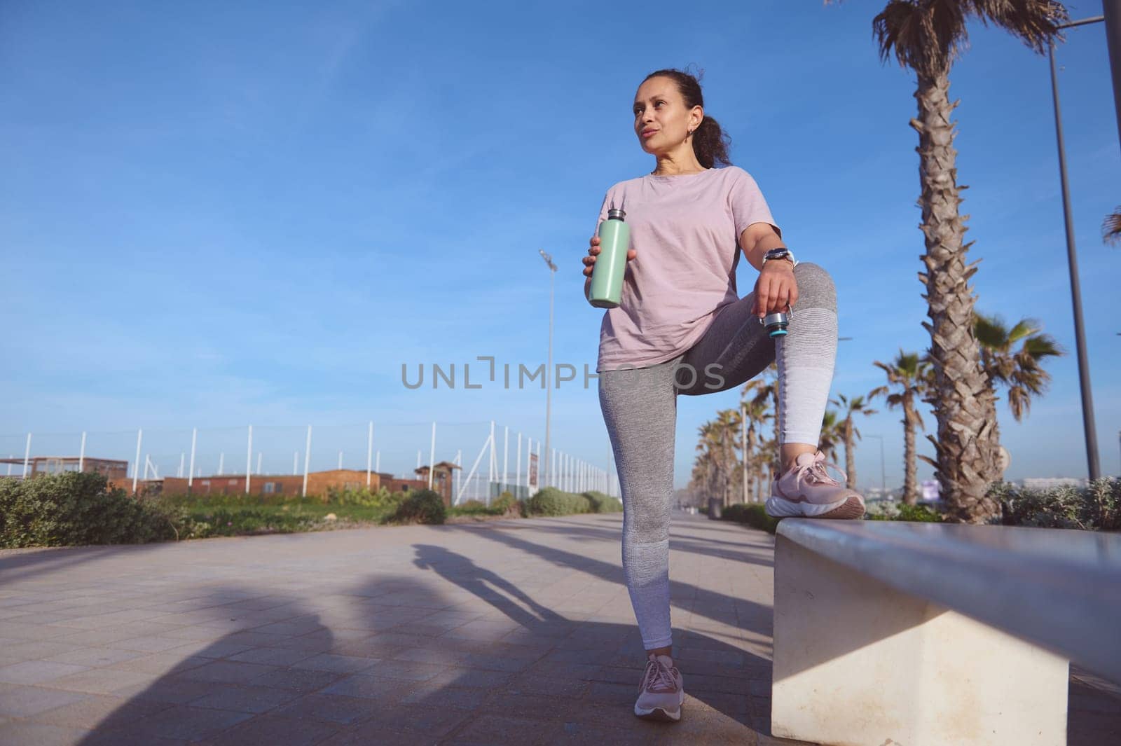 Full length Latina sportswoman relaxing after morning jog, drinking water from a steel bottle. People. Fitness. Sport by artgf