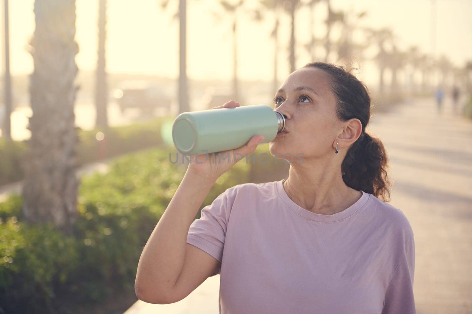 Curly haired Latina woman in purple t-shirt, drinking water from a steel bottle, standing on the treadmill and resting after jog at sunset. People. Active and healthy lifestyle. Sport. Cardio workout