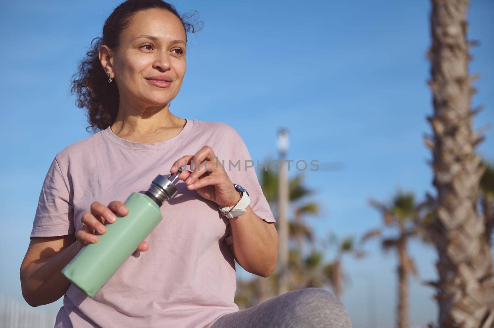 Authentic portrait of a multi ethnic pretty woman, wonderful female runner standing outside, holding water bottle. Wellness lady taking a break after running workout. Active people and sport concept