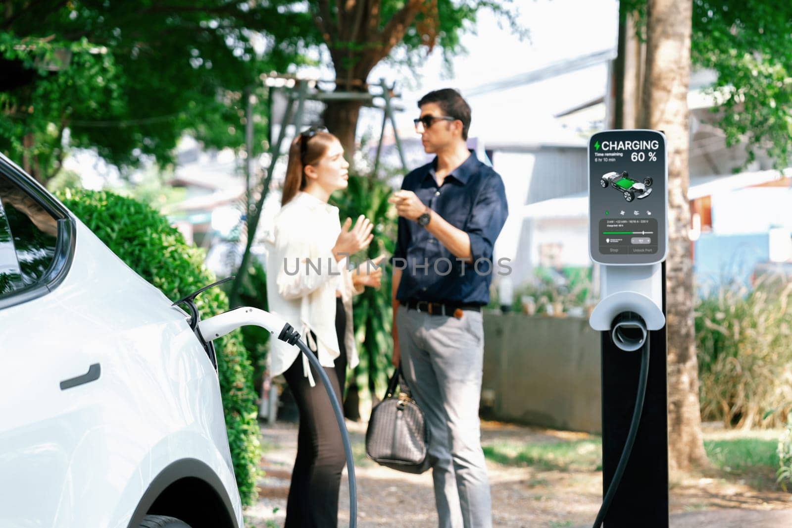 Young couple recharge electric car's battery from charging station in outdoor green city park in springtime. Rechargeable EV car for sustainable environmental friendly urban travel lifestyle.Expedient