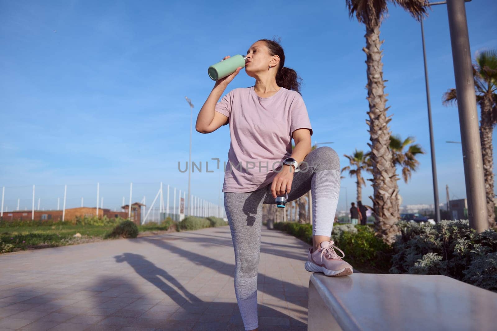 Attractive sporty girl taking break after outdoor workout or morning jog, standing by a stone bench and drinking water by artgf