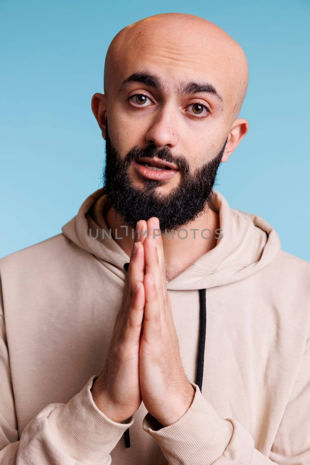 Young adult arab man with folded arms asking for help while looking at camera with pleading facial expression. Person pleasing, praying to god and making wish studio portrait