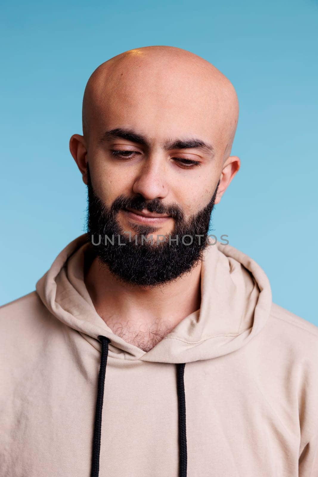 Young adult arab man looking down with thoughtful facial expression. Puzzled bald bearded person thinking, wearing casual beige hoodie and posing naturally on blue background