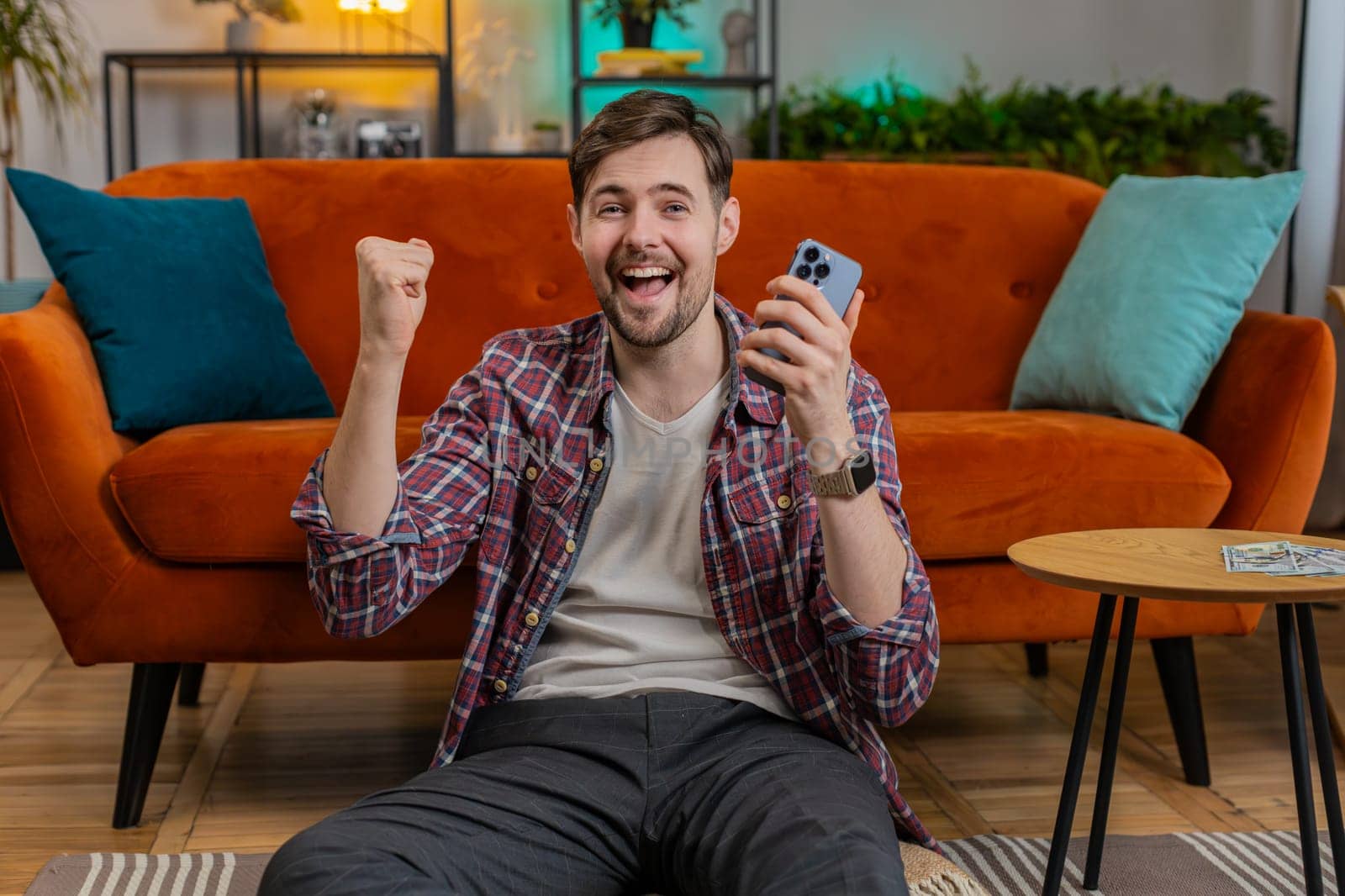 Surprised excited man winner holds smartphone reading good news amazed by online bet bid game lottery jackpot casino win. Happy Caucasian guy overjoyed by victory success shows thumbs up at home floor