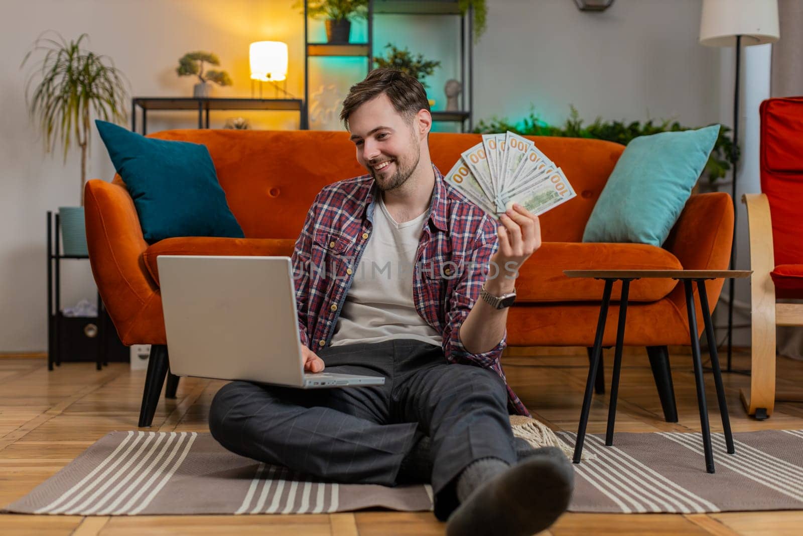 Planning budget. Rich happy Caucasian man counting money cash dollar using laptop calculate domestic bills at home. Young adult guy satisfied of income earnings, saves money for planned vacation