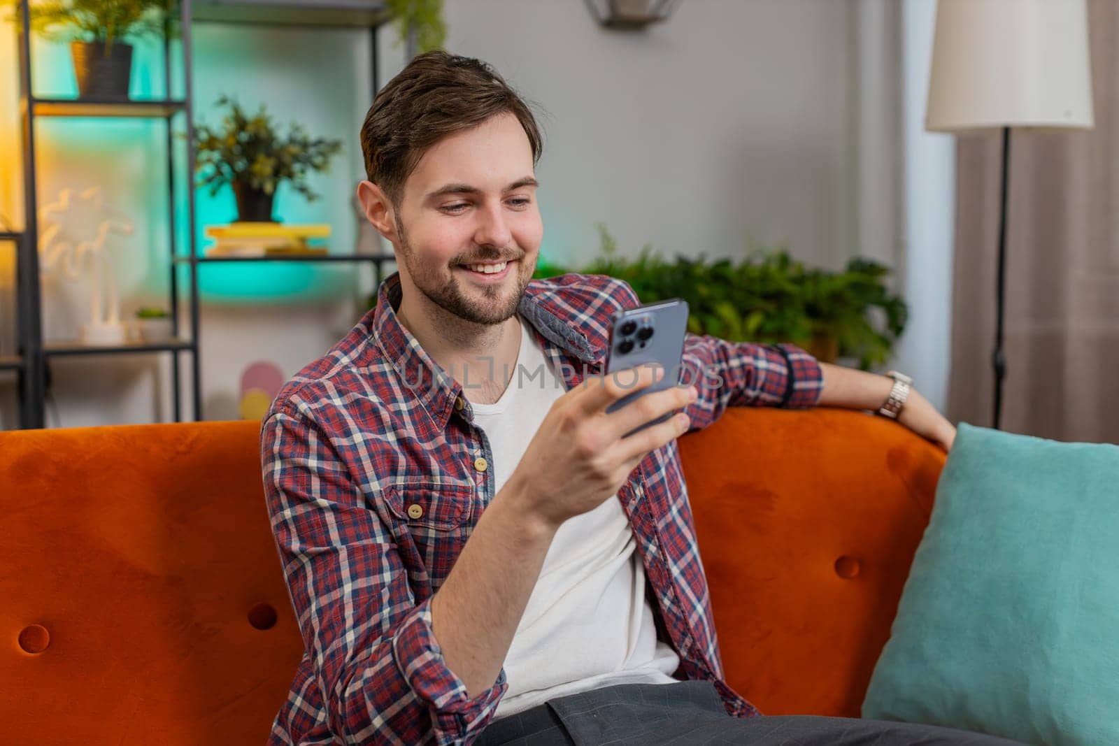 Happy smiling man in casual clothes thinking and text messaging on smartphone. Caucasian guy sitting on sofa and using mobile phone in living room at home. Male is watching online movie in apartment.