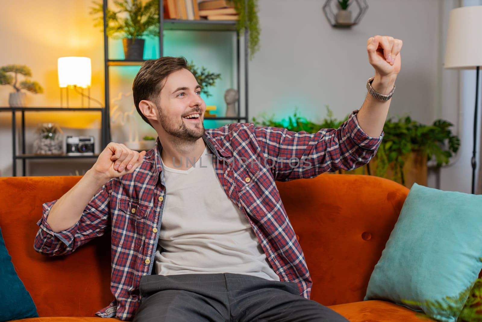 Energetic young man singing and dancing with hands while sitting on sofa in living room. Carefree Caucasian guy celebrate victory win enjoying favorite music having fun while staying alone at home.