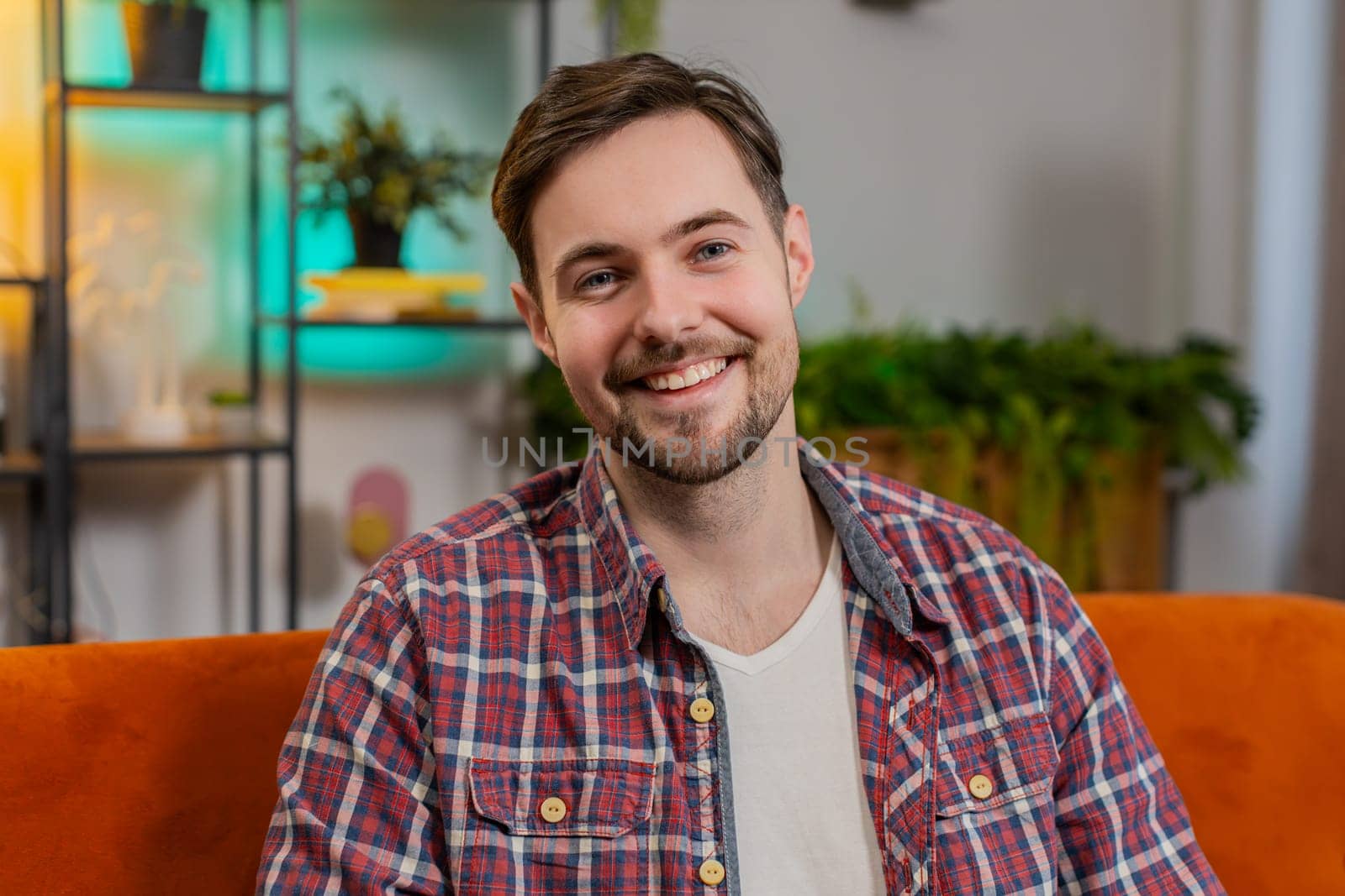 Portrait of happy Caucasian man sitting on sofa looking at camera and smiling at home. Young guy in plaid shirt having attractive appearance enjoying leisure time relaxing in living room in apartment