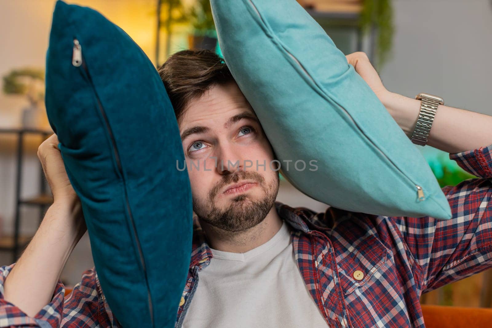 Irritated Caucasian tired man annoyed by noisy neighbors suffers from headache on sofa. Thin walls at home, flat without sound insulation, repair work concept. Guy covers ear with cushion pillows.