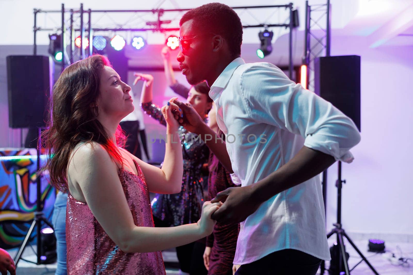 Romantic young couple holding hands and dancing together while clubbing. Diverse boyfriend and girlfriend partying and moving on dancefloor in nightclub illuminated with lights