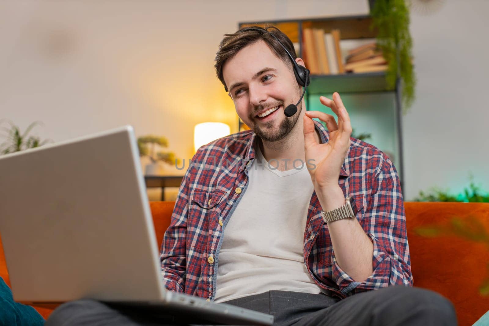 Happy excited business man helpline in plaid shirt talking to web camera headset making an online video conference call in distance. Caucasian freelancer guy uses laptop on sofa in living room at home