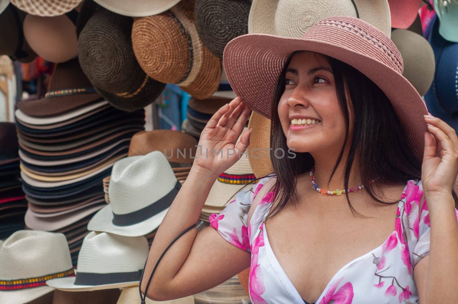 A cheerful woman tries on various hats, surrounded by a colorful selection at an outdoor market stall on a sunny day. by Raulmartin