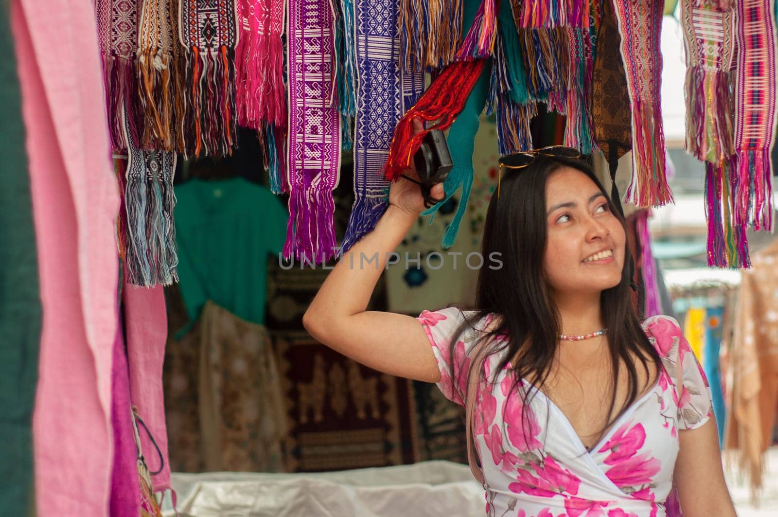 A smiling young woman in a floral dress browses through vibrant, hanging scarves at an outdoor market stall. High quality photo