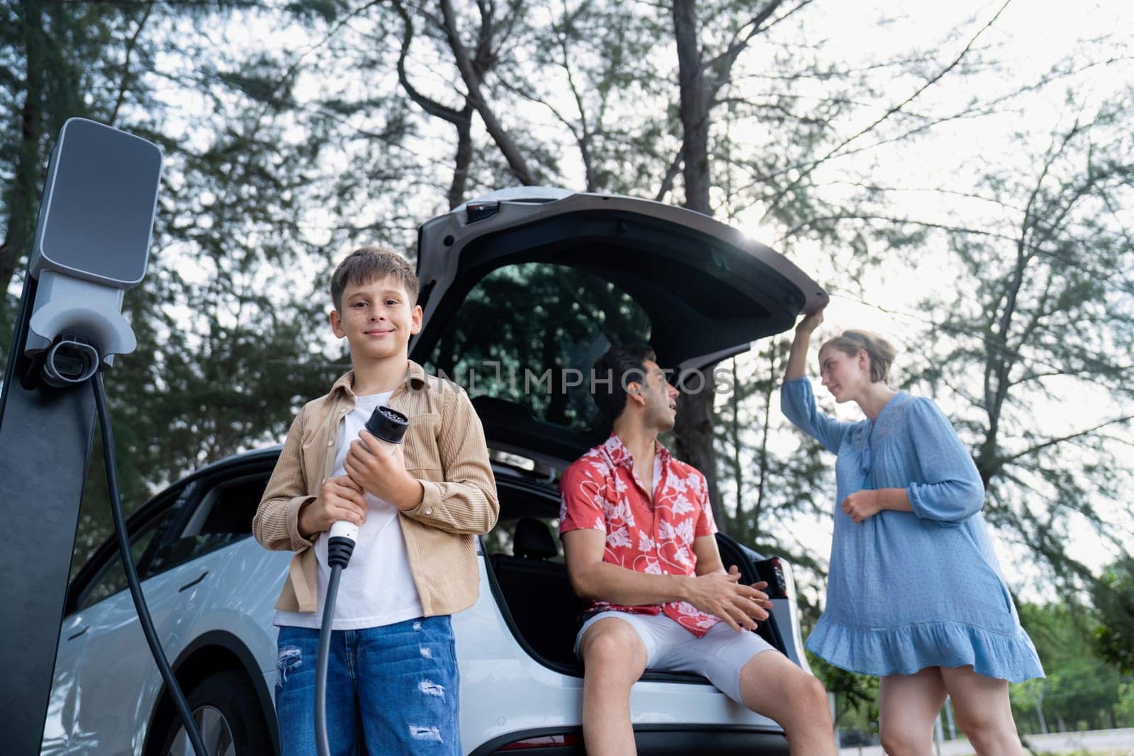 Little boy holding EV charger and point at camera with his family sitting on the trunk in background. Road trip travel with alternative energy charging station for eco-friendly car concept. Perpetual