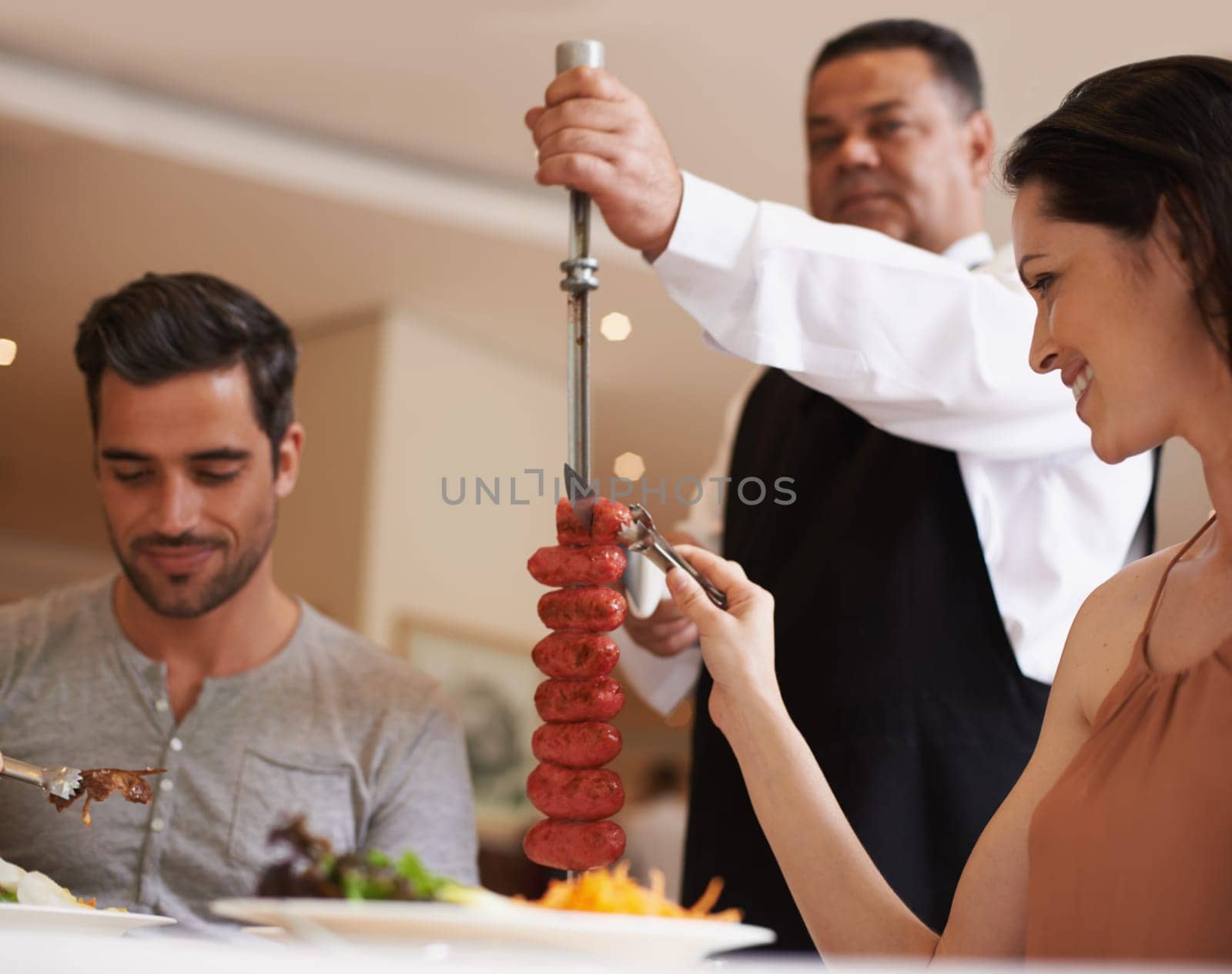Couple, waiter and service for lunch in restaurant with meat, happiness and fine dining for anniversary or honeymoon. Man, woman and employee with sausage on skewer, healthy meal and table in London.