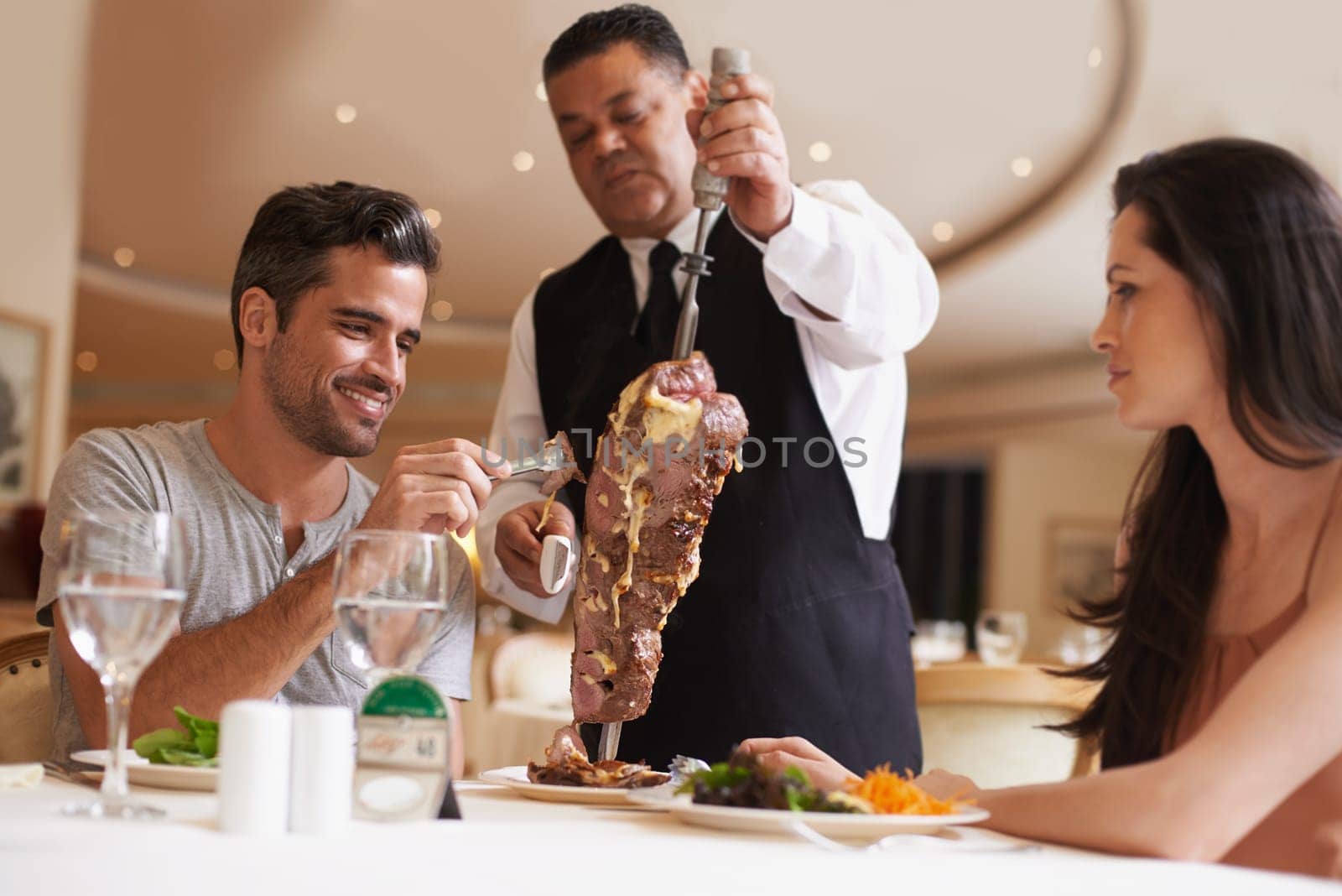 Couple, waiter and service for dinner in restaurant with meat, happiness and fine dining for anniversary or honeymoon. Man, woman and employee with steak on skewer, healthy meal and table in London.