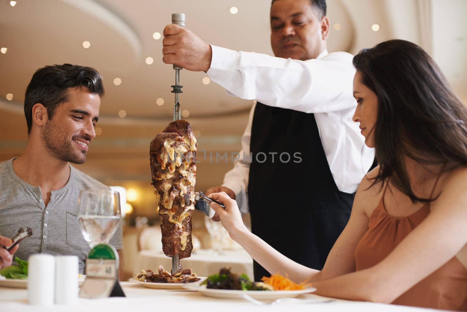 Couple, waiter and service for lunch in restaurant with meat, happiness and fine dining for anniversary or honeymoon. Man, woman and employee with steak on skewer, healthy meal and table in London.
