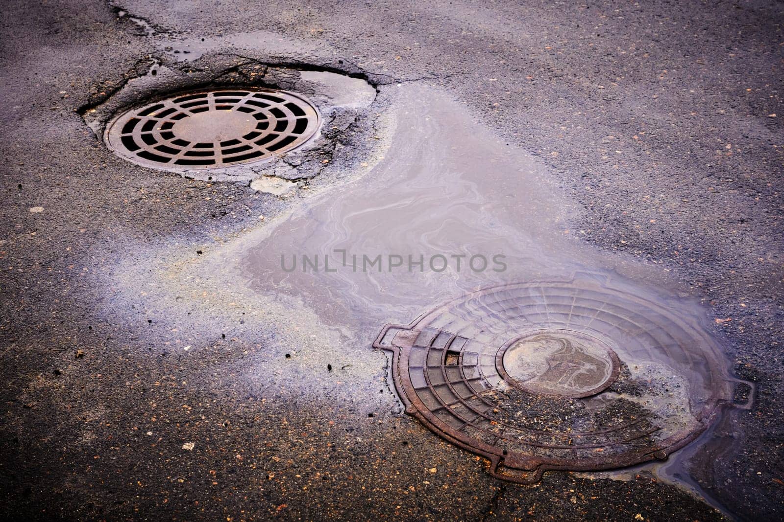 An oil slick against the background of an asphalt road flows into a storm drain against the background of a sewer grate and the lid of a closed well