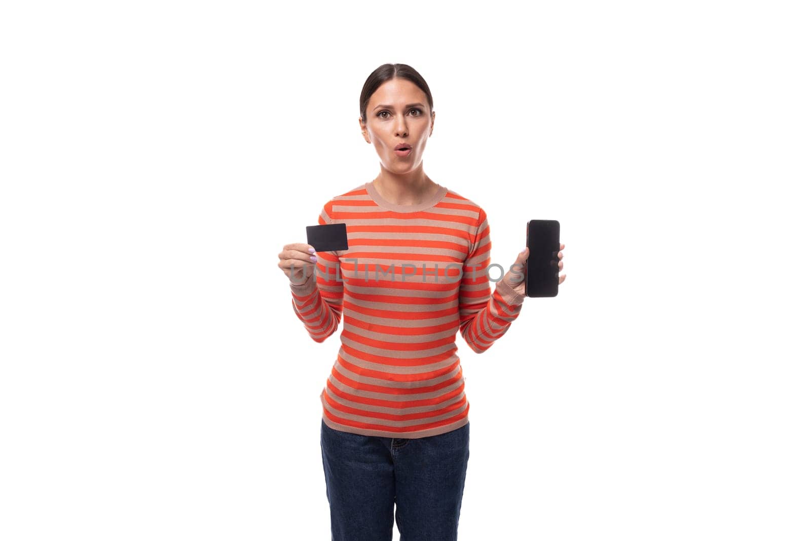A young pretty brunette woman dressed in a striped orange blouse with surprise shows a plastic card and a smartphone.