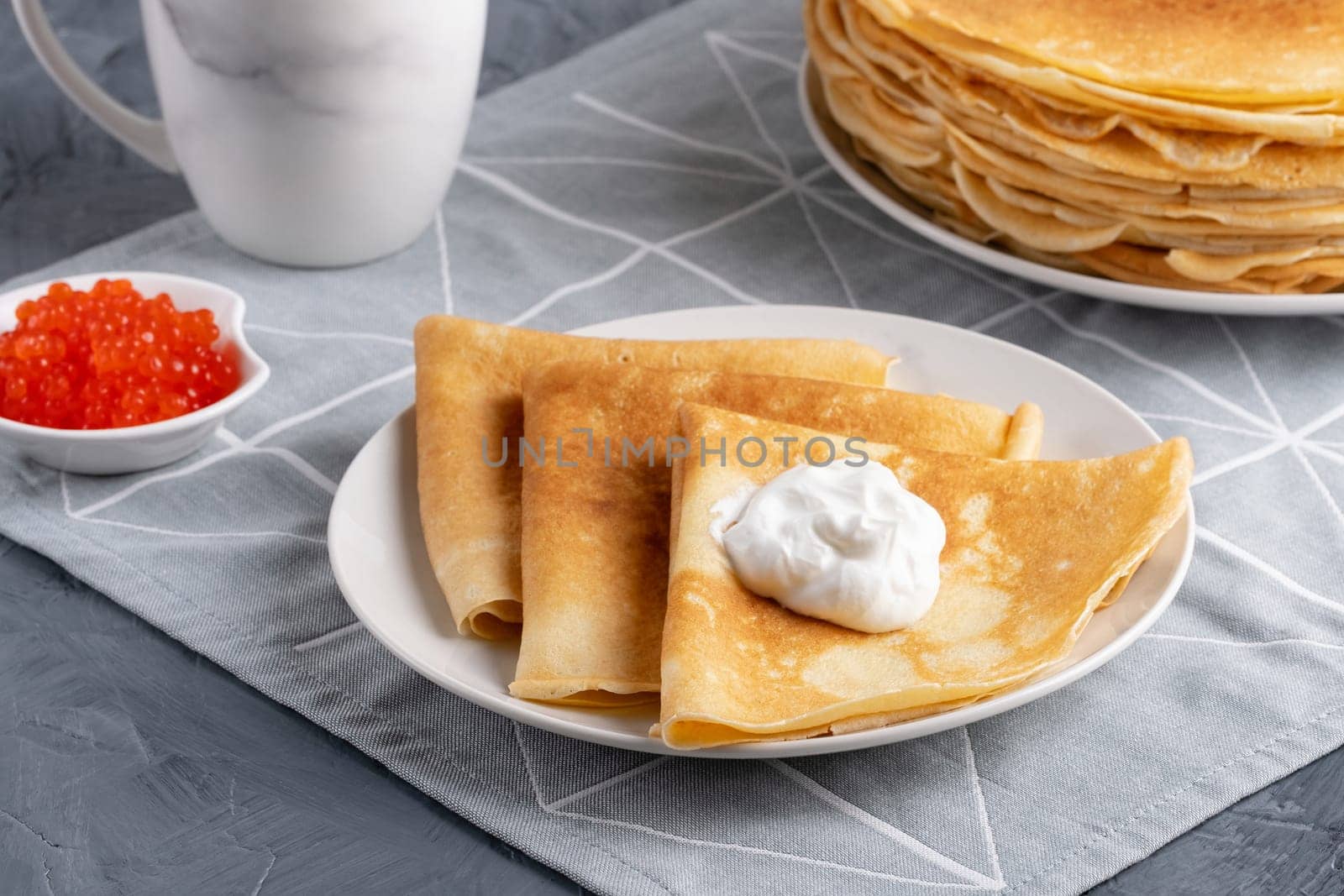 Pancakes with sour cream and red caviar. Close-up of pancakes stacked in a plate on a gray background.