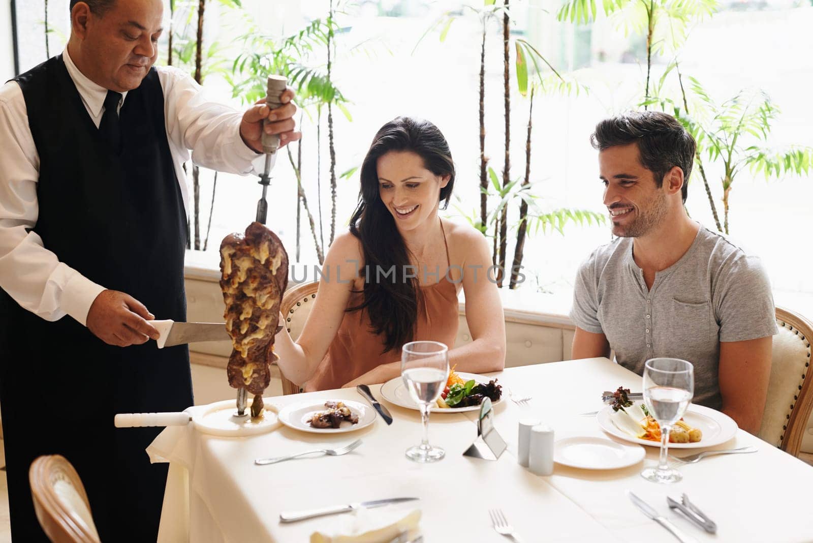 Happy couple, date and waiter with meat kebab for serving, fine dinning or romantic dinner at table. Young man and woman enjoying chef cutting skewer of food for meal, eating or service at restaurant.