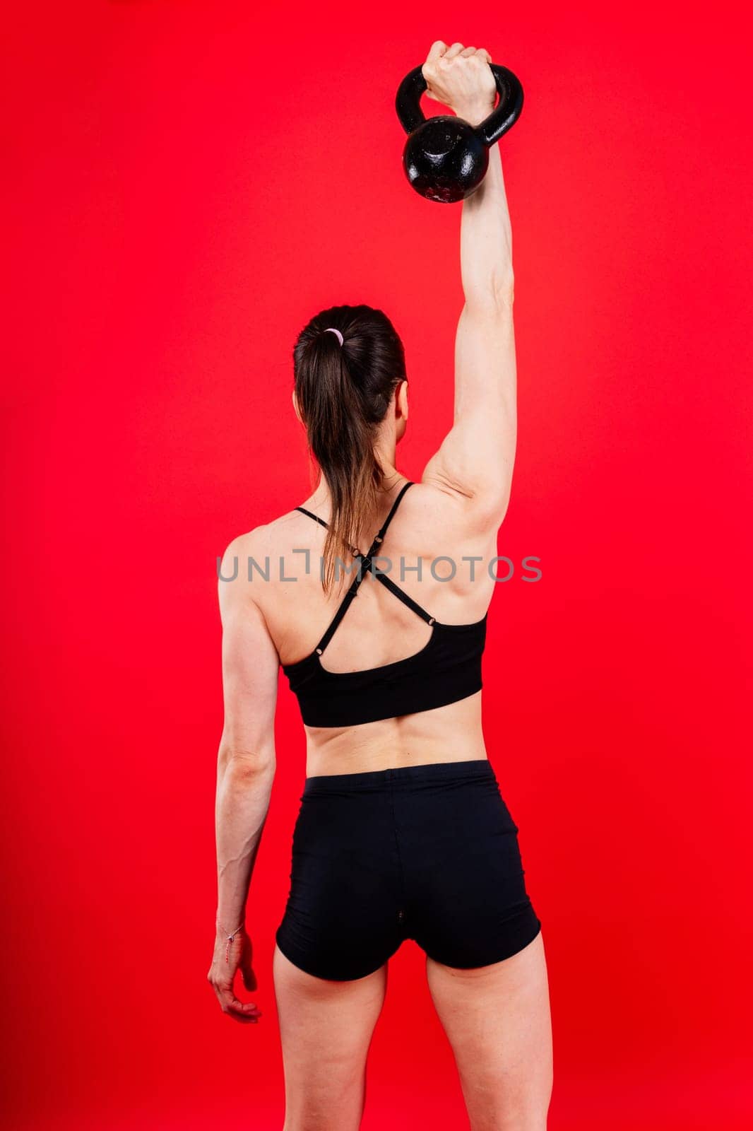 Woman in sportswear doing crossfit workout with kettle bell on dark yellow red background. by Zelenin