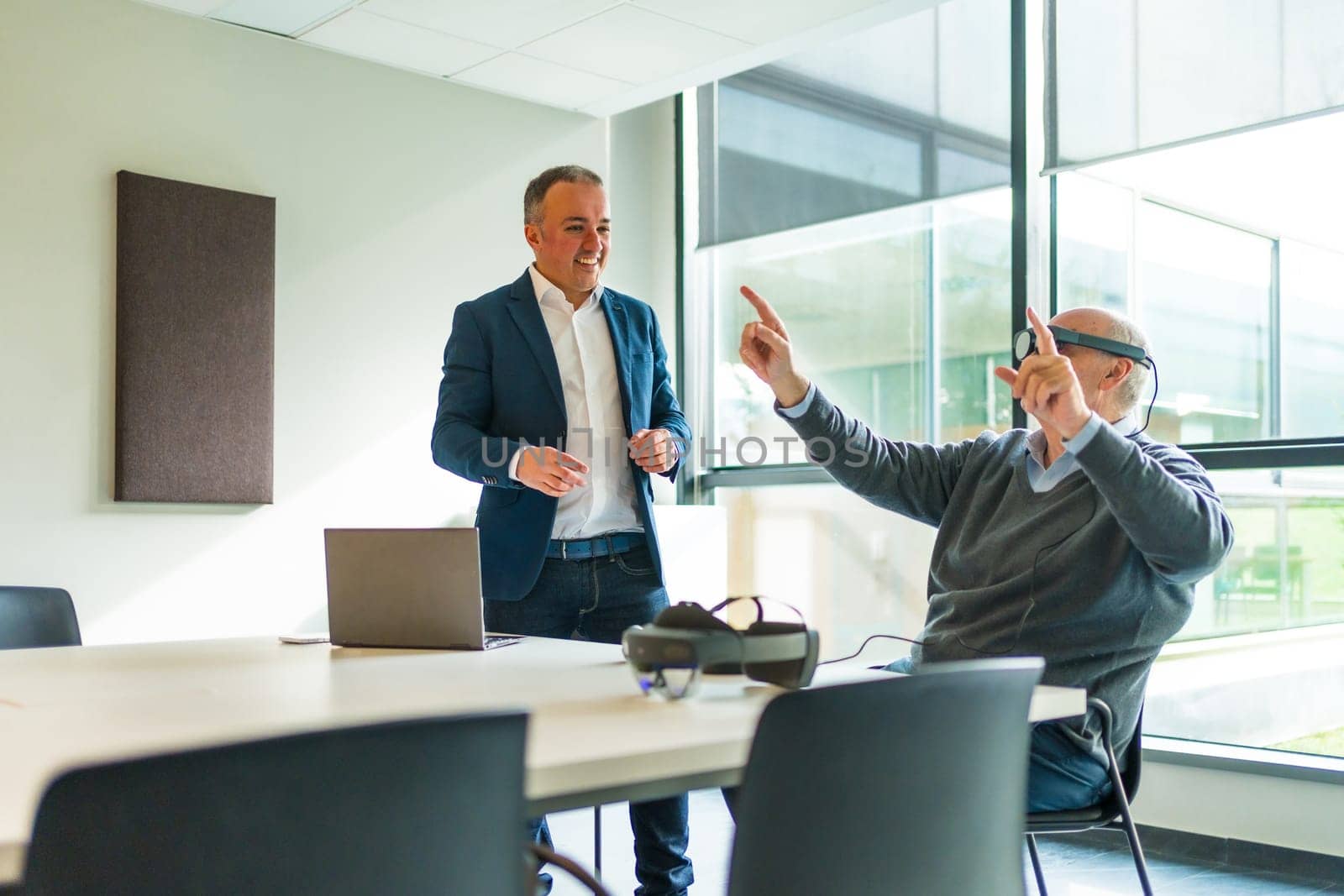 Aged manager sitting and using VR goggles next to a employee in a office