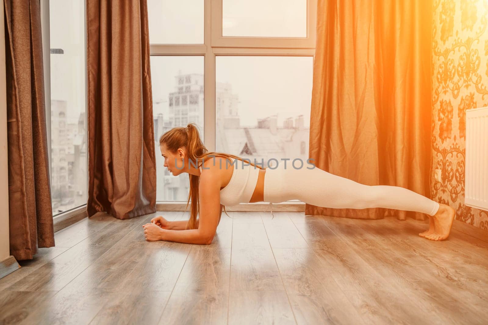 Young attractive athletic woman in white sportswear doing push ups or pushups on gym mat at home, practicing yoga plank pose, exercising indoors, healthy active lifestyle concept by Matiunina