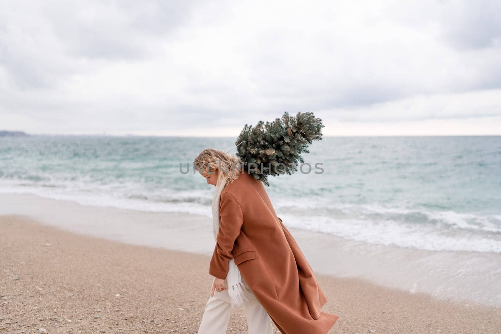 Blond woman Christmas sea. Christmas portrait of a happy woman walking along the beach and holding a Christmas tree on her shoulder. She is wearing a brown coat and a white suit