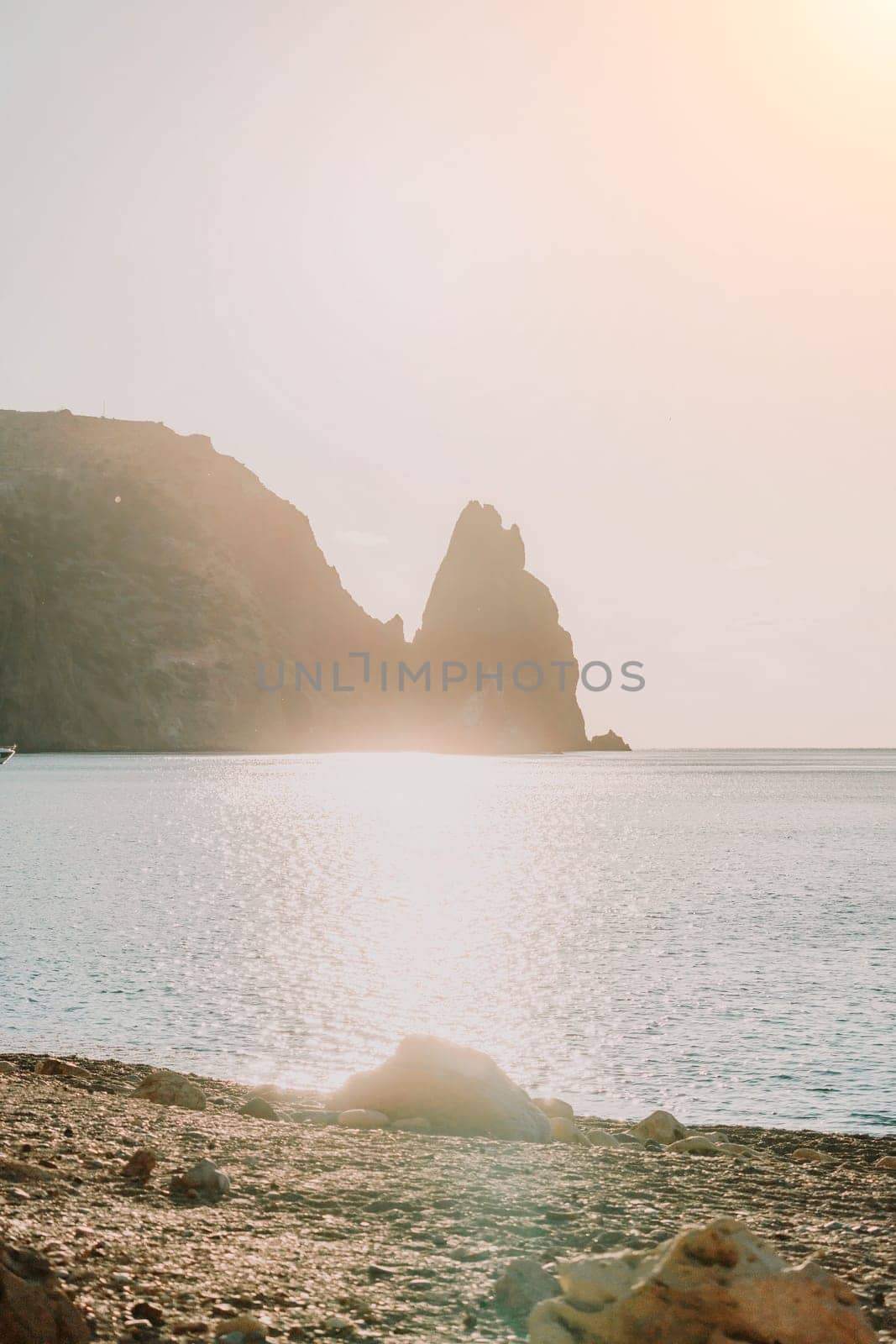 A red burning sunset with the silhouette of a cliff over the sea. Abstract nature summer or spring ocean sea background. Small waves on golden warm water surface with bokeh lights from sun.