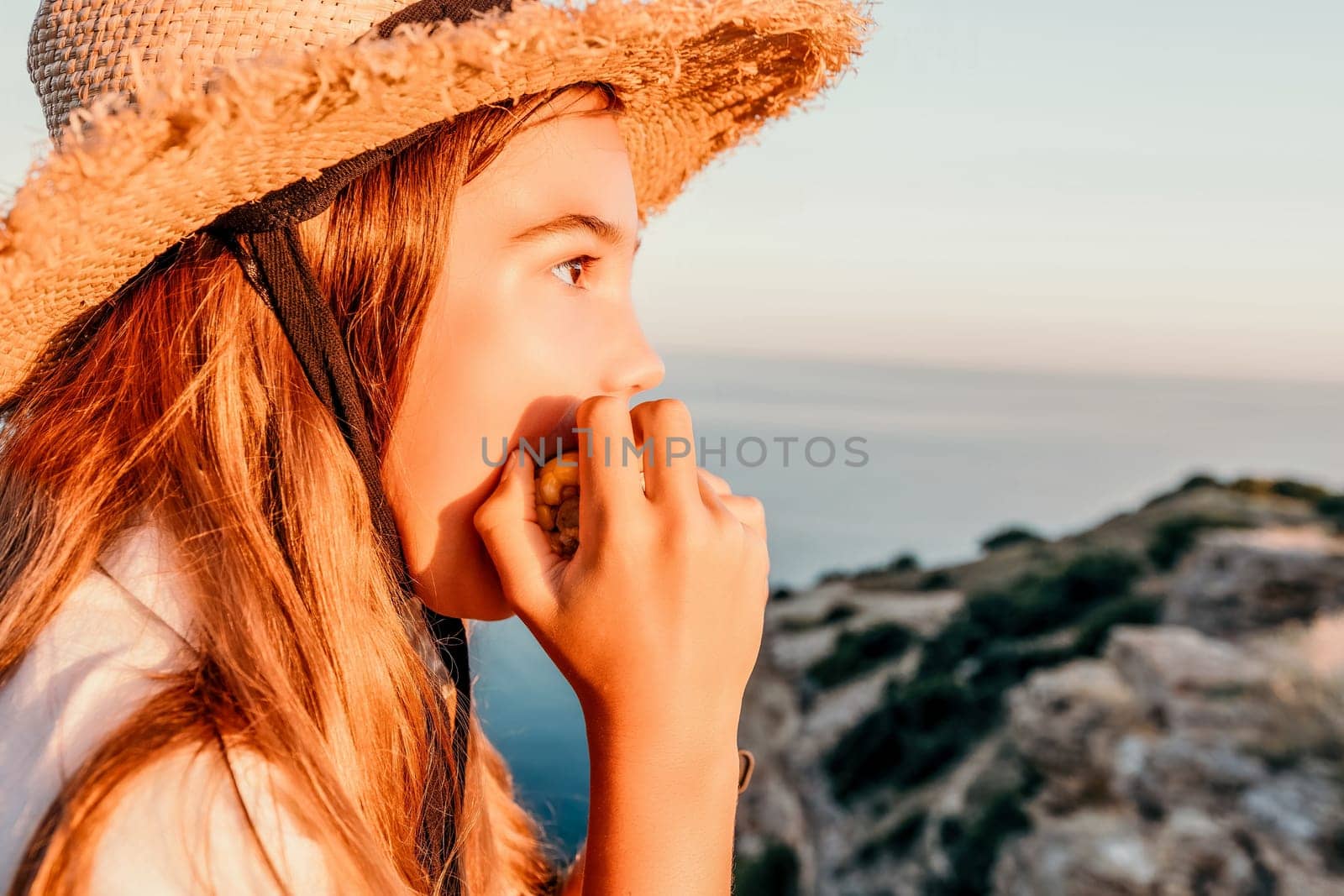Happy girl eating corn. Summer snacking on the sea. Portrait of young beautiful girl in straw hat eating grilled corn while sitting by the sea on sunset time. Close up. Selective focus by panophotograph