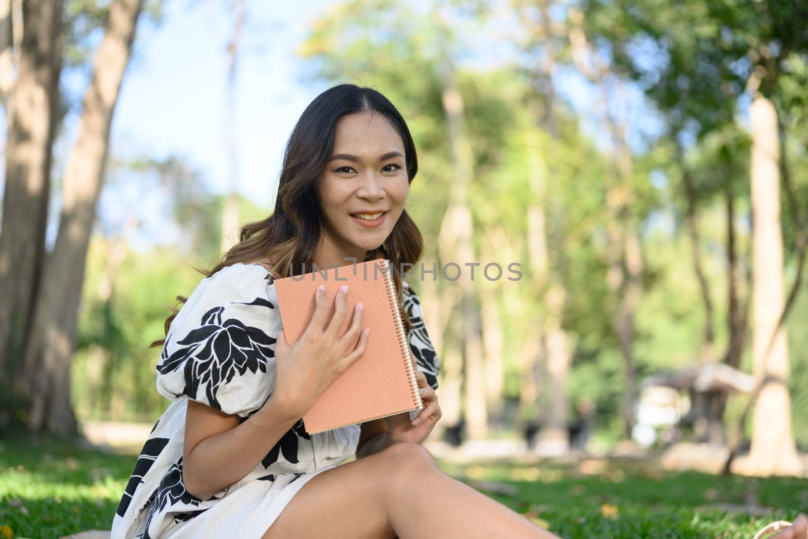 Pleased young woman reading favorite novel while sitting on green grass at public park by prathanchorruangsak