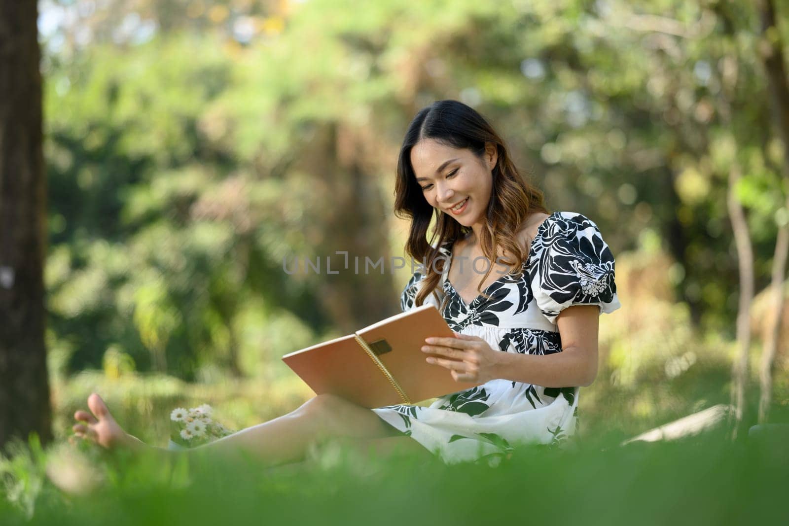 Relaxed young Asian woman reading interesting book and enjoying free time at green park by prathanchorruangsak