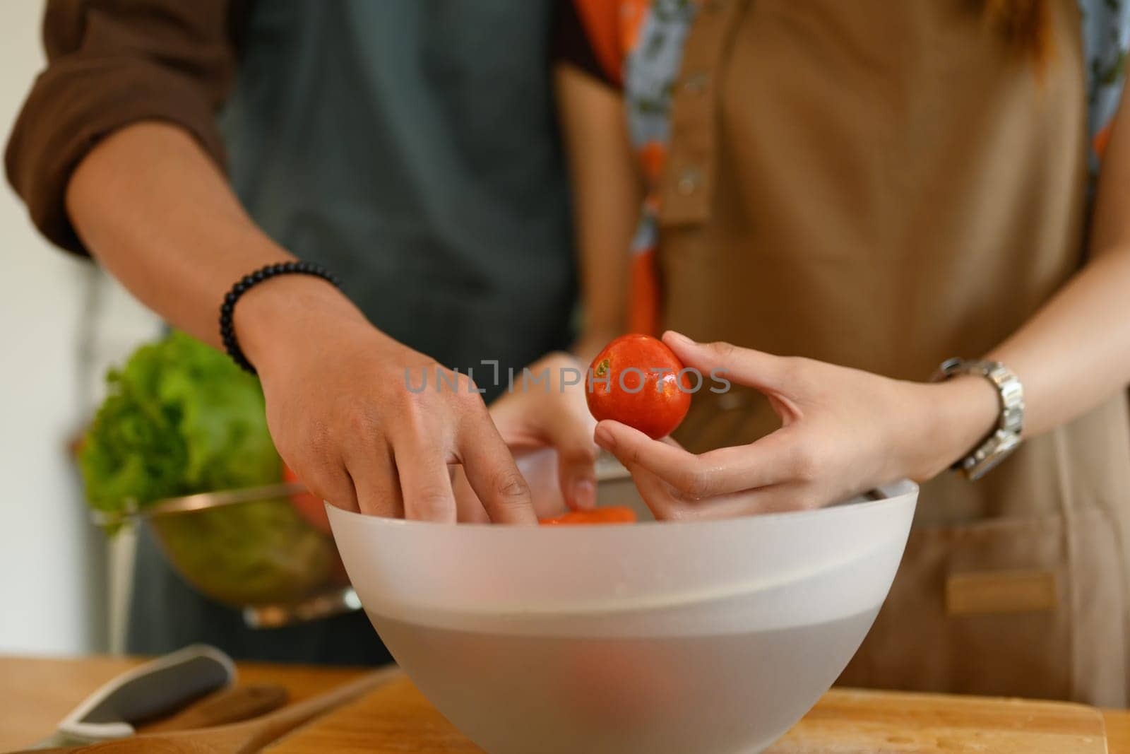 Young couple in aprons preparing healthy salad with organic vegetables on kitchen table.