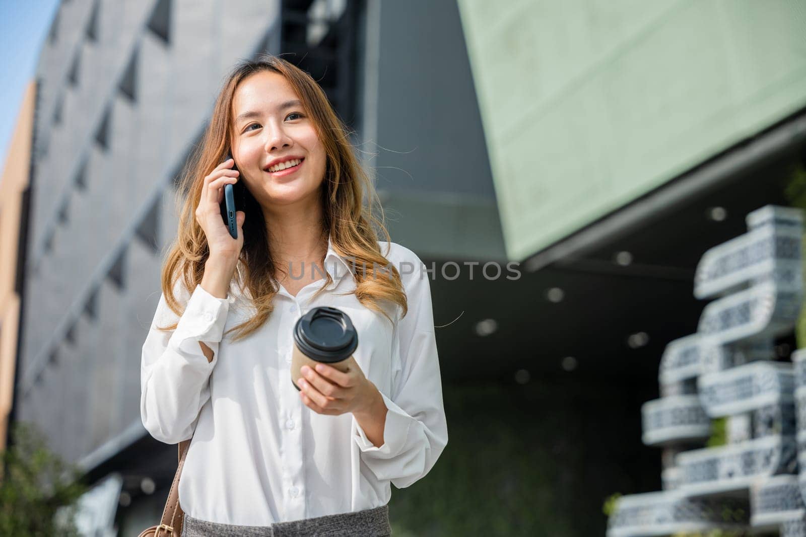 Beautiful woman walking outside in street and calling talking on mobile phone outdoors near office, portrait lifestyle businesswoman with bag cell to customer while holding coffee paper cup takeaway