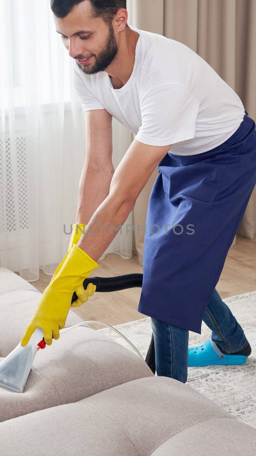 Close-up of housekeeper holding modern washing vacuum cleaner and cleaning dirty sofa with professionally detergent. Professional springclean at home concept by Mariakray